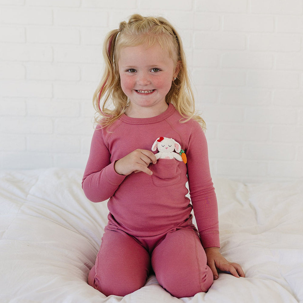 A young blonde girl with pigtails sits smiling on a white bed, wearing a matching dusty rose long-sleeve pajama top and pants. She is holding a small white plush toy with colorful accents.