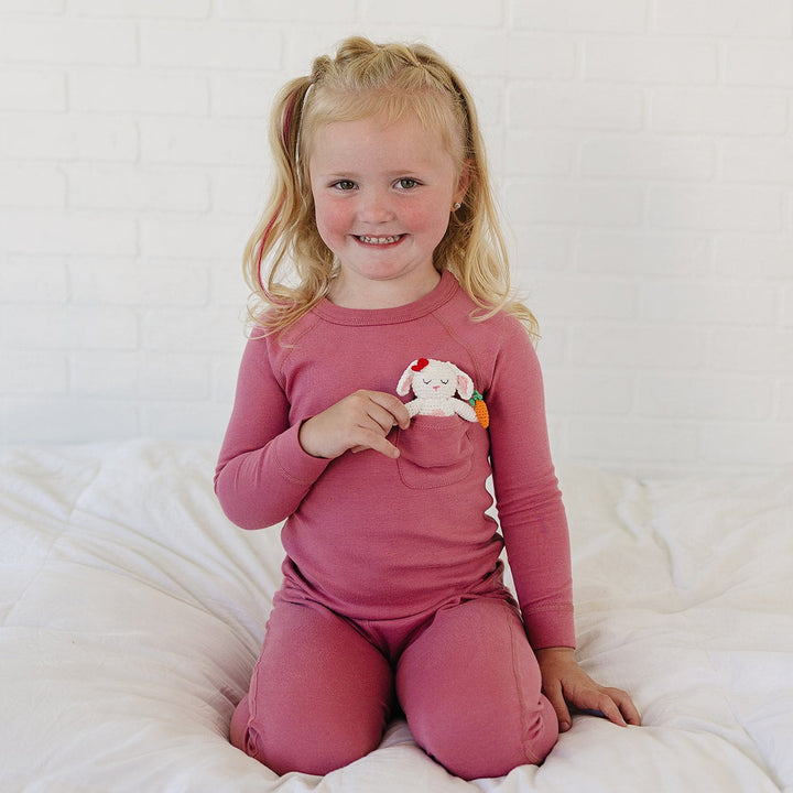A young blonde girl with pigtails sits smiling on a white bed, wearing a matching dusty rose long-sleeve pajama top and pants. She is holding a small white plush toy with colorful accents.