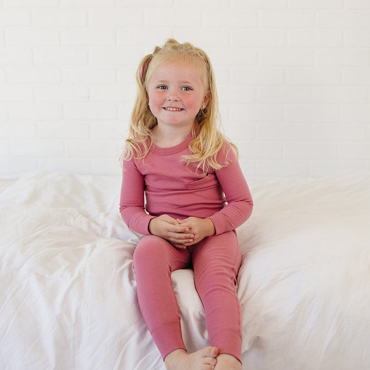 A young blonde girl sitting on a white bed wearing a matching dusty rose pink long-sleeve top and leggings set.