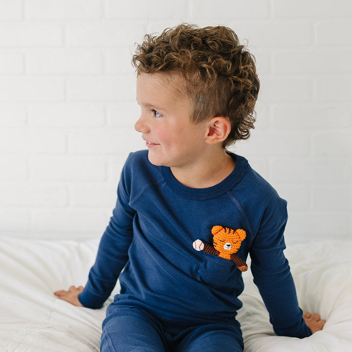 A young boy with curly brown hair wearing a navy blue long-sleeve pajama set with an orange tiger patch on the pocket, sitting on a white bed against a white brick wall.