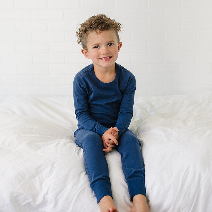 A young boy with curly light brown hair sits on a white bed wearing a navy blue long-sleeve pajama set against a white brick wall background.