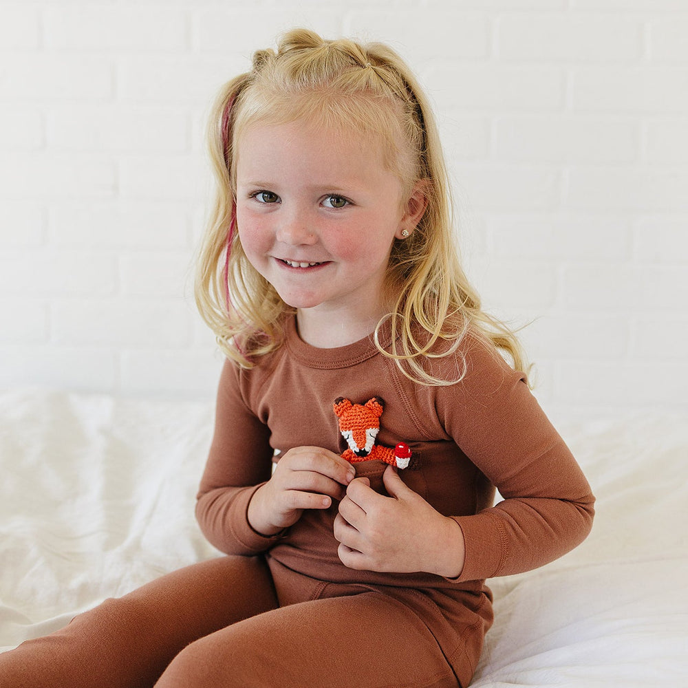 A young blonde girl with pigtails sits on a white bed wearing a matching brown long-sleeve top and pants set. She is holding a small orange and white plush fox toy against her chest.