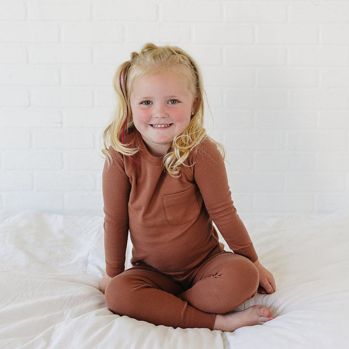 A young blonde girl sitting cross-legged on a white bed, wearing a matching rust-brown long-sleeve top and leggings set against a white brick wall background.