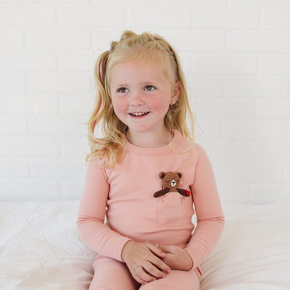 A young blonde girl with pigtails sits smiling on a white bed, wearing a light pink long-sleeve pajama top with a small embroidered brown bear on the chest and matching pink pajama pants.