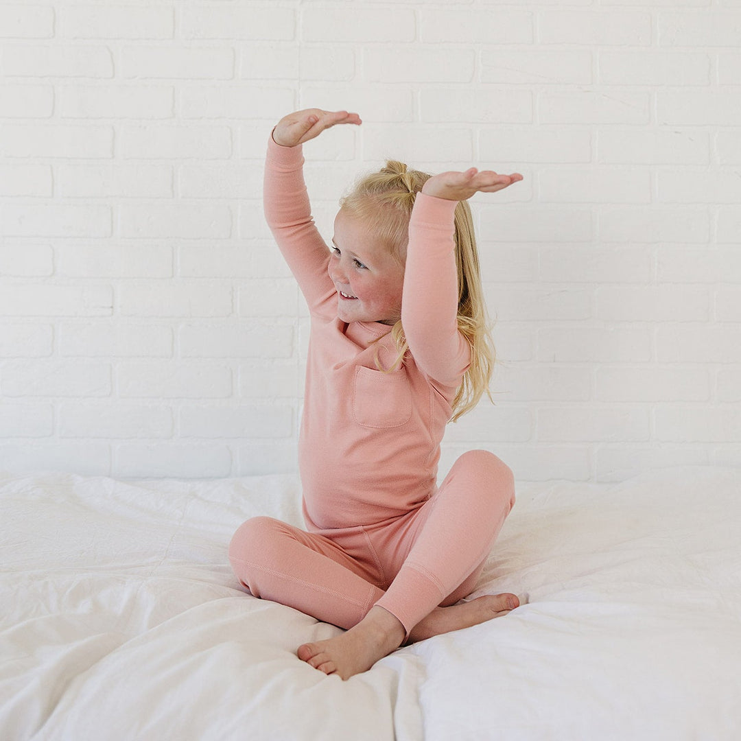 A young girl with blonde hair sitting on a white bed, wearing a matching dusty pink long-sleeve top and leggings set. She is stretching her arms overhead against a white brick wall background.
