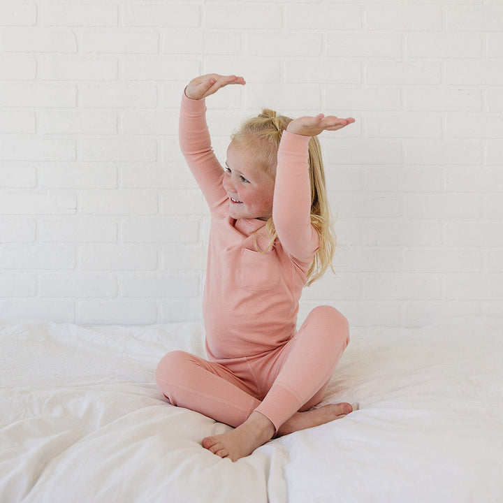 A young girl with blonde hair sitting on a white bed, wearing a matching dusty pink long-sleeve top and leggings set. She is stretching her arms overhead against a white brick wall background.