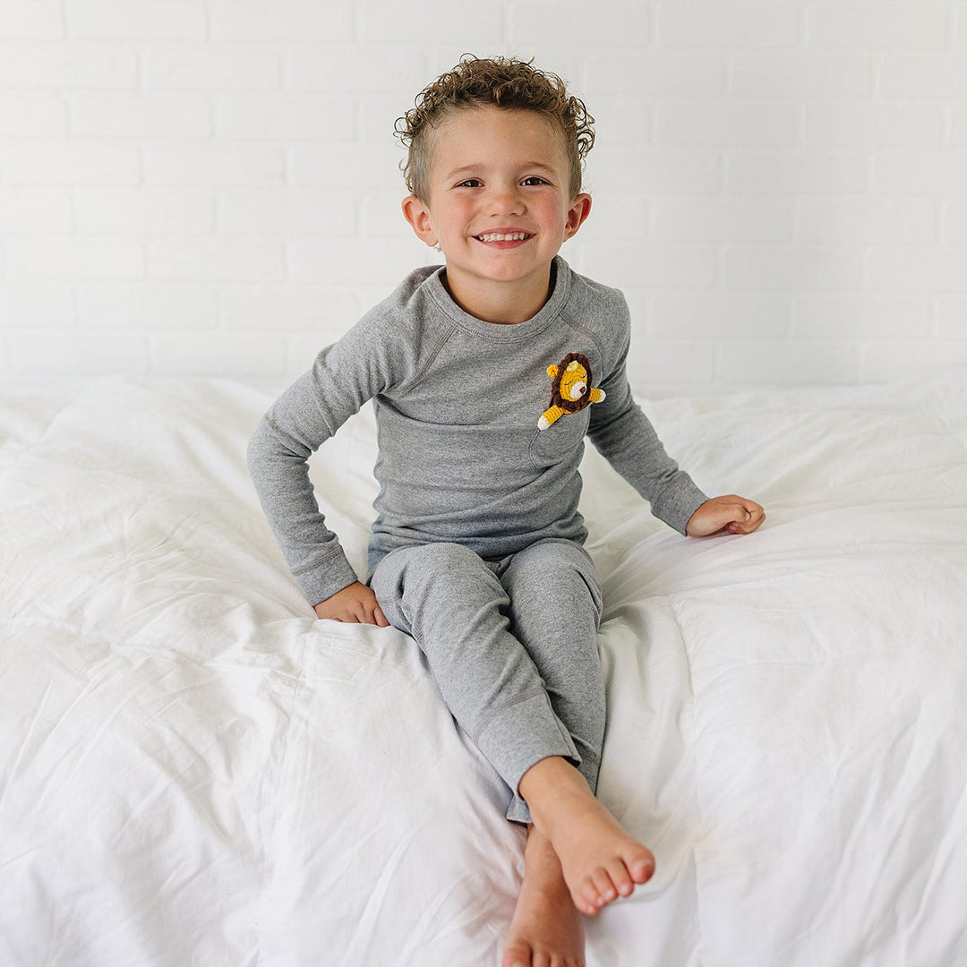 A smiling young boy with curly hair sits on a white bed wearing a gray long-sleeve pajama top and matching pants, with a small yellow lion patch on the chest.