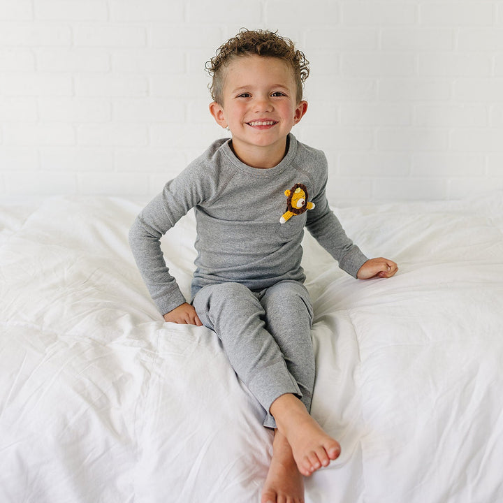 A smiling young boy with curly hair sits on a white bed wearing a gray long-sleeve pajama top and matching pants, with a small yellow lion patch on the chest.