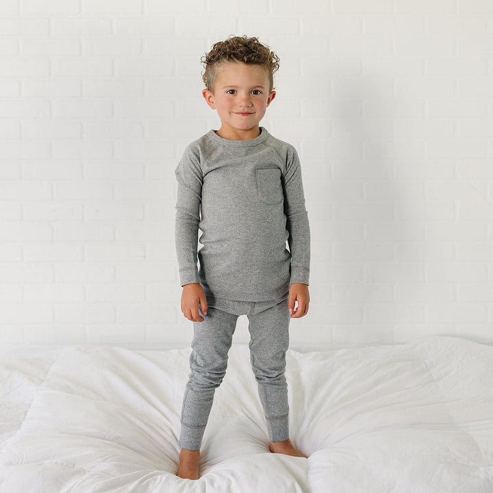 A young boy with curly hair stands barefoot on a white bed wearing a heather gray long-sleeve shirt and matching gray pants against a white brick wall background.