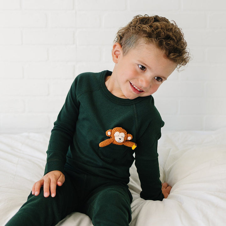 A toddler boy wearing a dark green long-sleeve pajama top and matching pants, featuring a crocheted monkey applique on the chest, sitting on a white bed against a white brick wall.