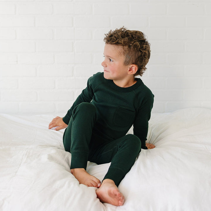 A young boy with curly brown hair sits on a white bed wearing a dark forest green long-sleeve pajama set against a white brick wall background.