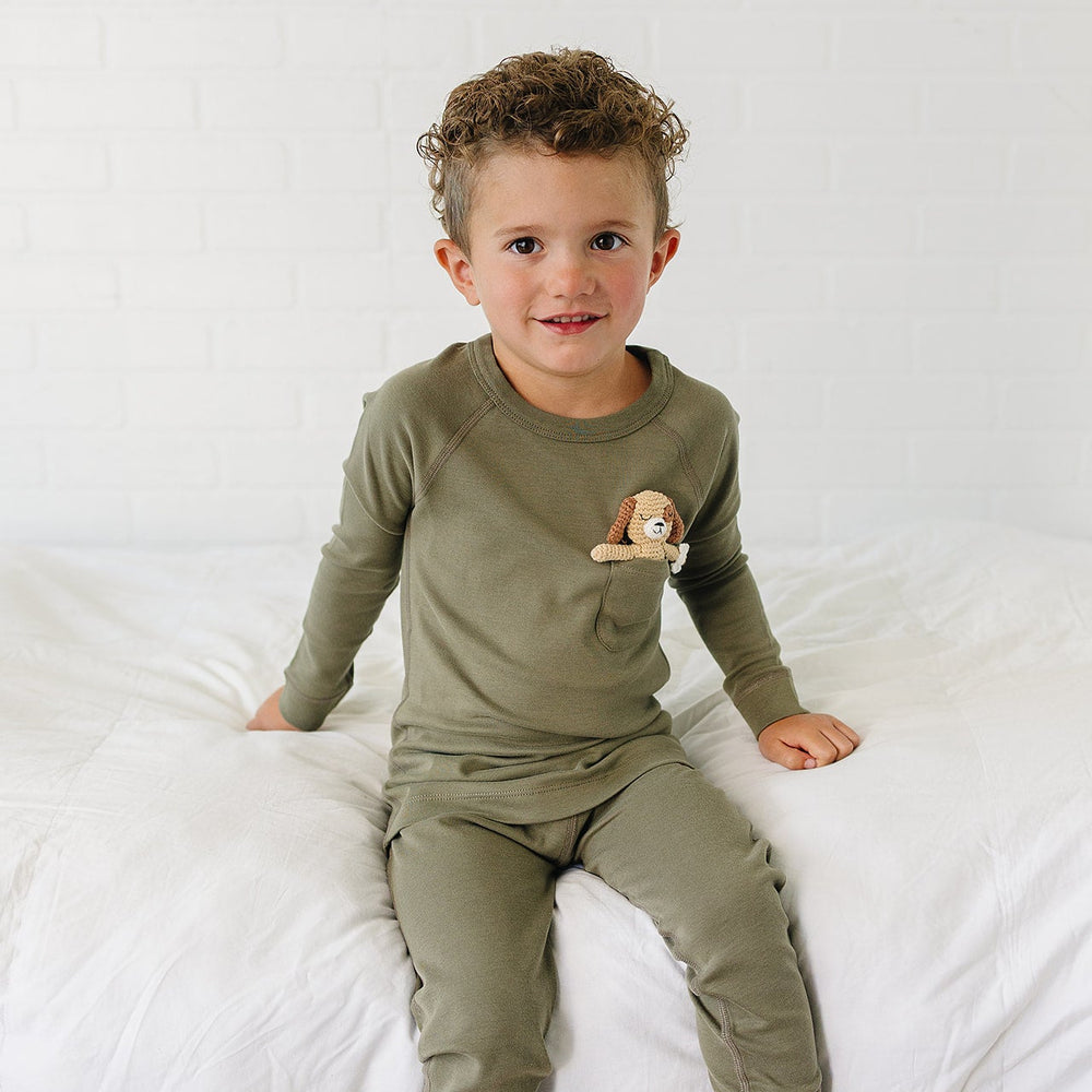 A toddler boy wearing an olive green long-sleeve pajama top and matching pants, sitting on a white bed against a white brick wall. A small plush dog is tucked into the chest pocket of the pajama top.