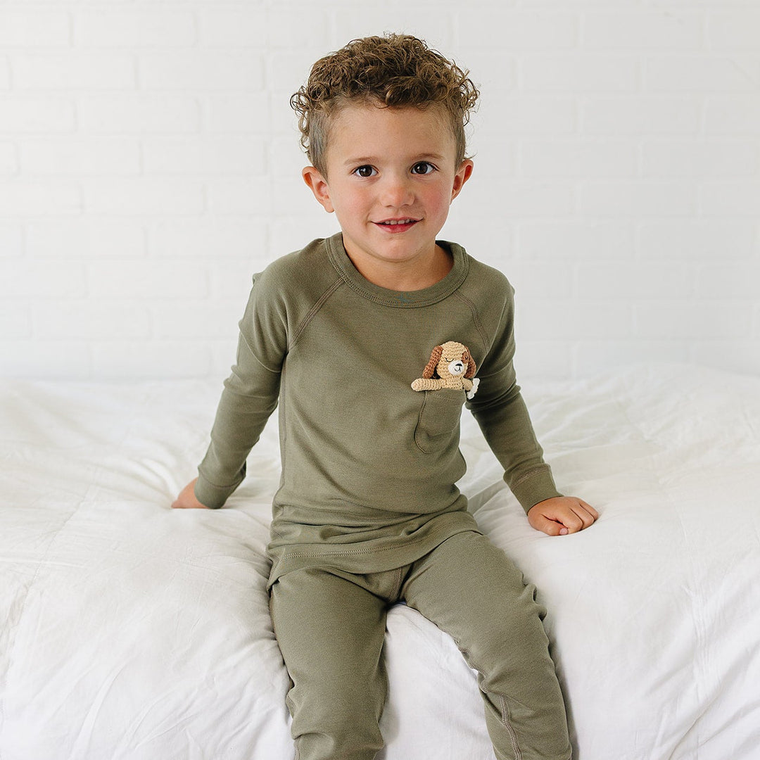 A toddler boy wearing an olive green long-sleeve pajama top and matching pants, sitting on a white bed against a white brick wall. A small plush dog is tucked into the chest pocket of the pajama top.