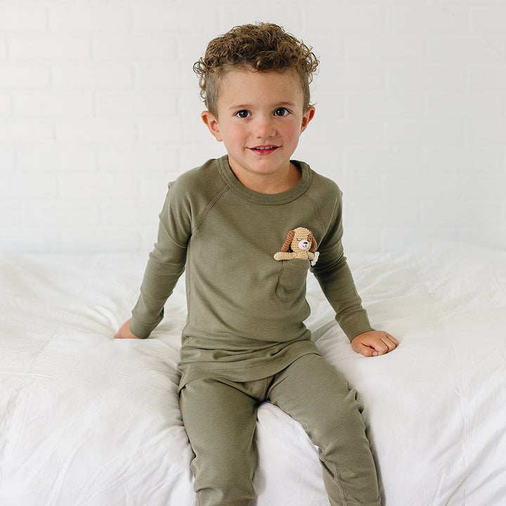 A toddler boy wearing an olive green long-sleeve pajama top and matching pants, sitting on a white bed against a white brick wall. A small plush dog is tucked into the chest pocket of the pajama top.