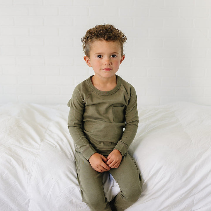 A young boy with curly brown hair sits on a white bed wearing an olive green long-sleeve crewneck shirt and matching green pants against a white brick wall background.