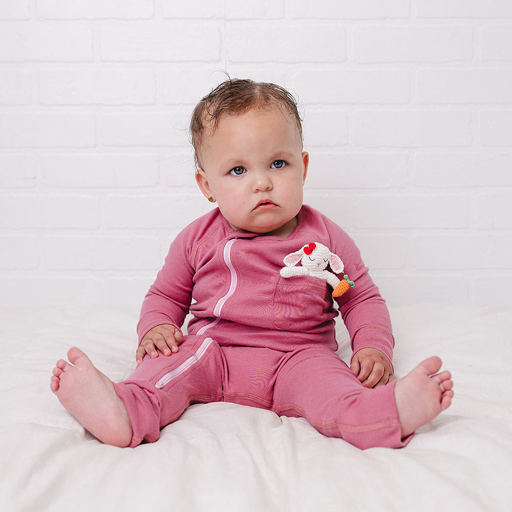 A baby sitting on a white bed against a white brick wall, wearing a pink long-sleeve romper and a small bunny on the chest.