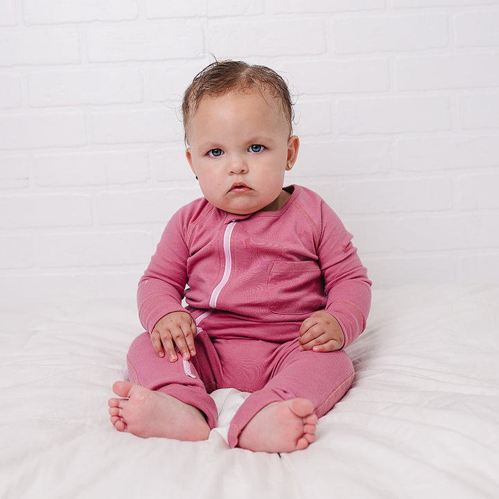 A baby sitting on a white bed against a white brick wall, wearing a pink long-sleeve footless pajama top with a light pink zipper.