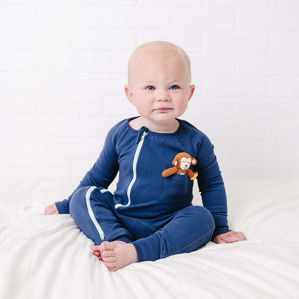 A baby sitting on a white bed wearing a navy blue zip-up one-piece pajama with white side stripes and a small monkey patch, set against a white brick wall.