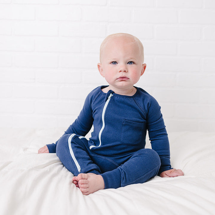 A baby sitting cross-legged on a white bed against a white brick wall, wearing a navy blue long-sleeve zip-up sleeper with white piping details.