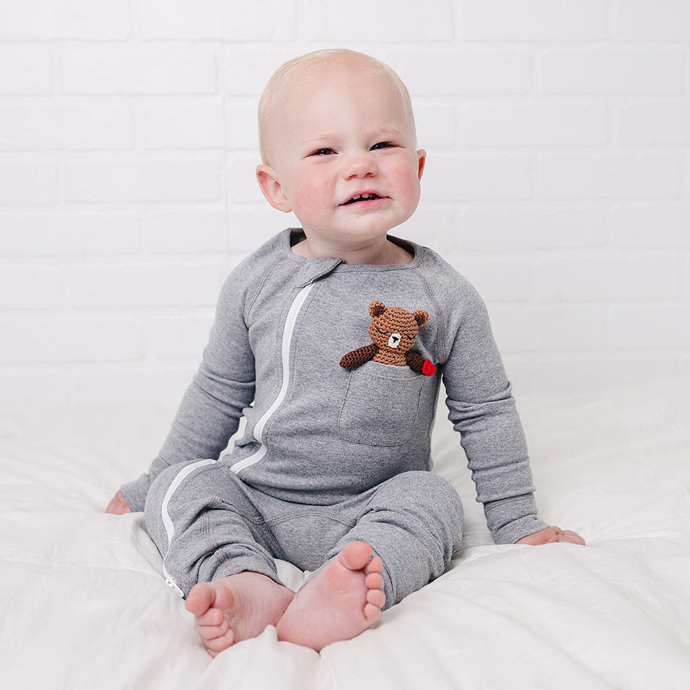 A baby sitting on a white bed wearing a gray long-sleeve zip-up onesie with a brown bear applique on the chest, set against a white brick wall background.