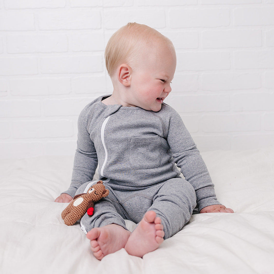 A baby sitting on a white bed wearing a gray long-sleeve one-piece pajama with a white zipper, holding a small brown toy against a white brick wall background.