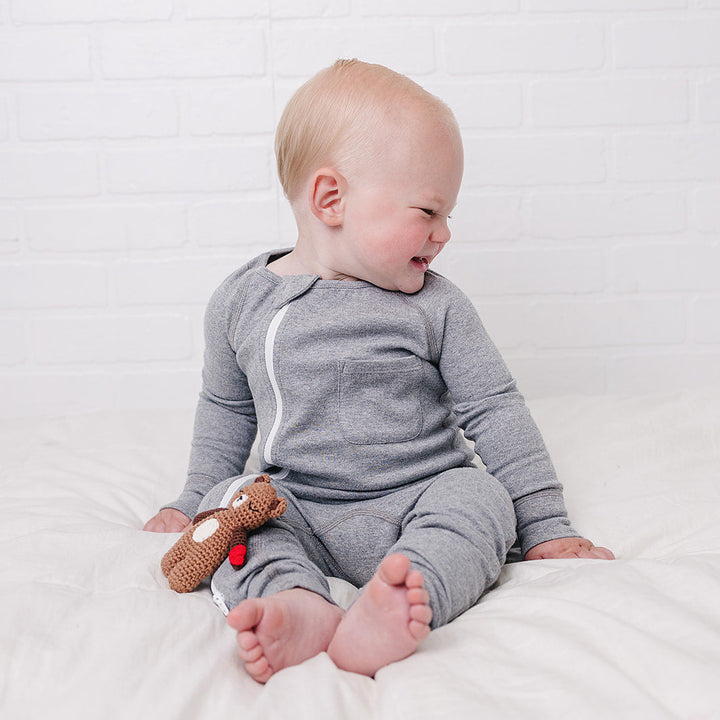 A baby sitting on a white bed wearing a gray long-sleeve one-piece pajama with a white zipper, holding a small brown toy against a white brick wall background.