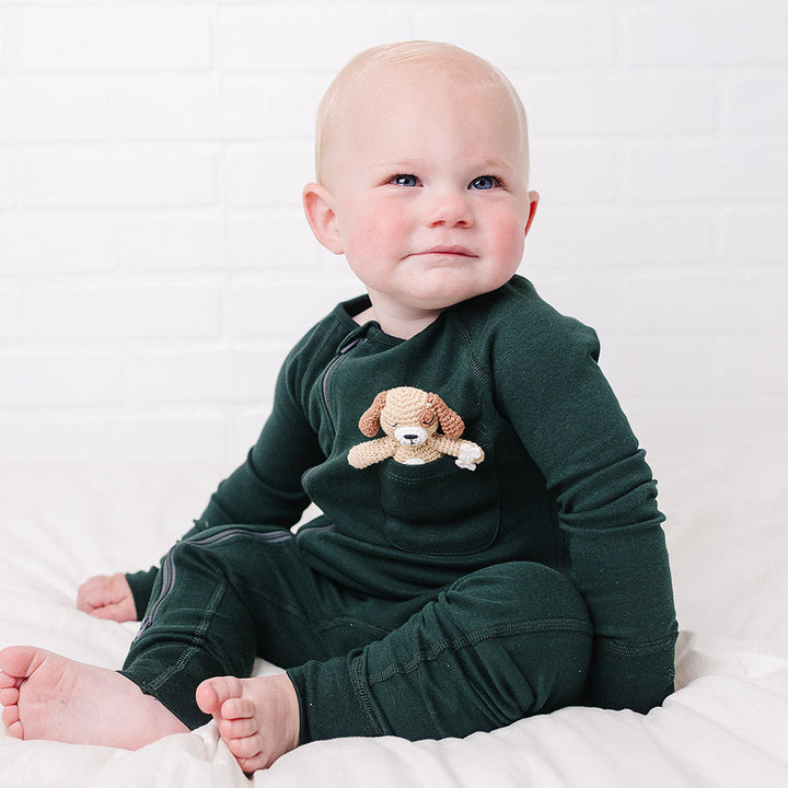 A baby sitting on a white bed wearing a dark green long-sleeve pajama set with a small teddy bear applique on the chest, set against a white brick wall background.