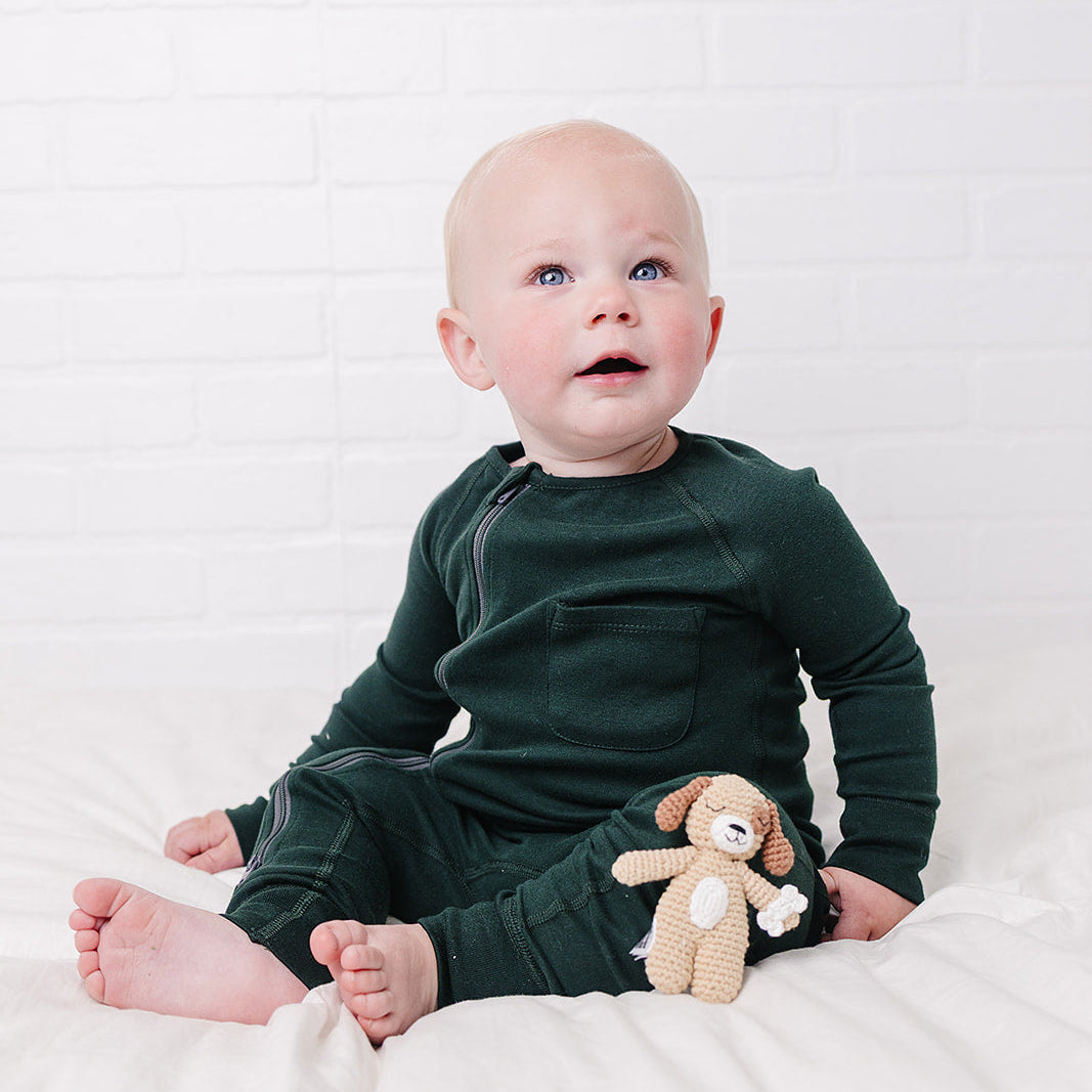 A baby sitting on a white bed wearing a dark forest green long-sleeve one-piece outfit and holding a small beige plush toy against a white brick wall background.